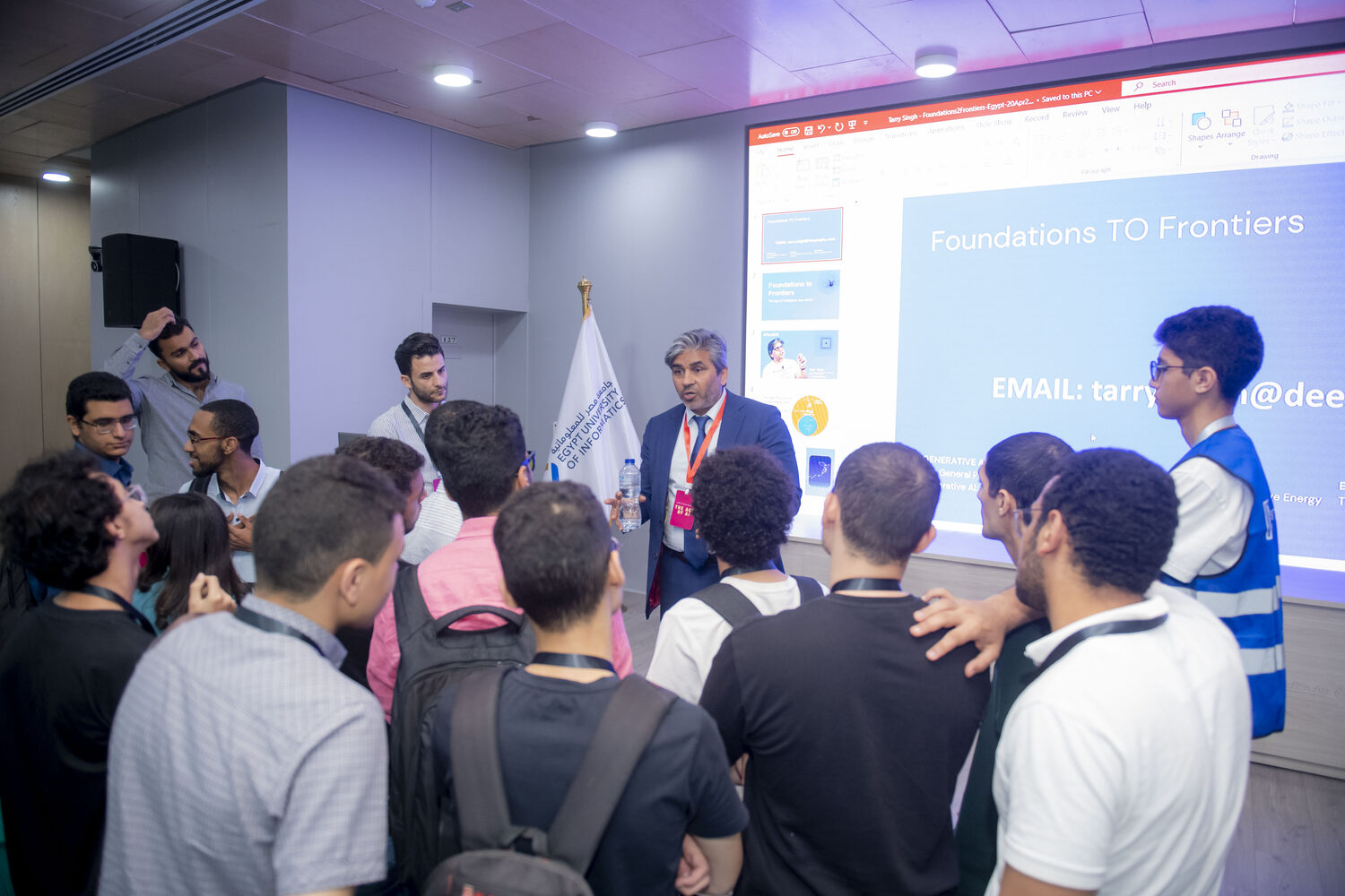 Students crowding the front of the room around Tarry Singh after the session, the Foundations TO Frontiers deck still on screen.
