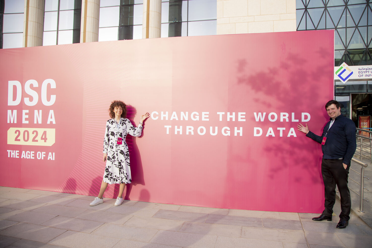 Two delegates with conference lanyards beside the DSC MENA 2024 Change the World Through Data wall outside the university.