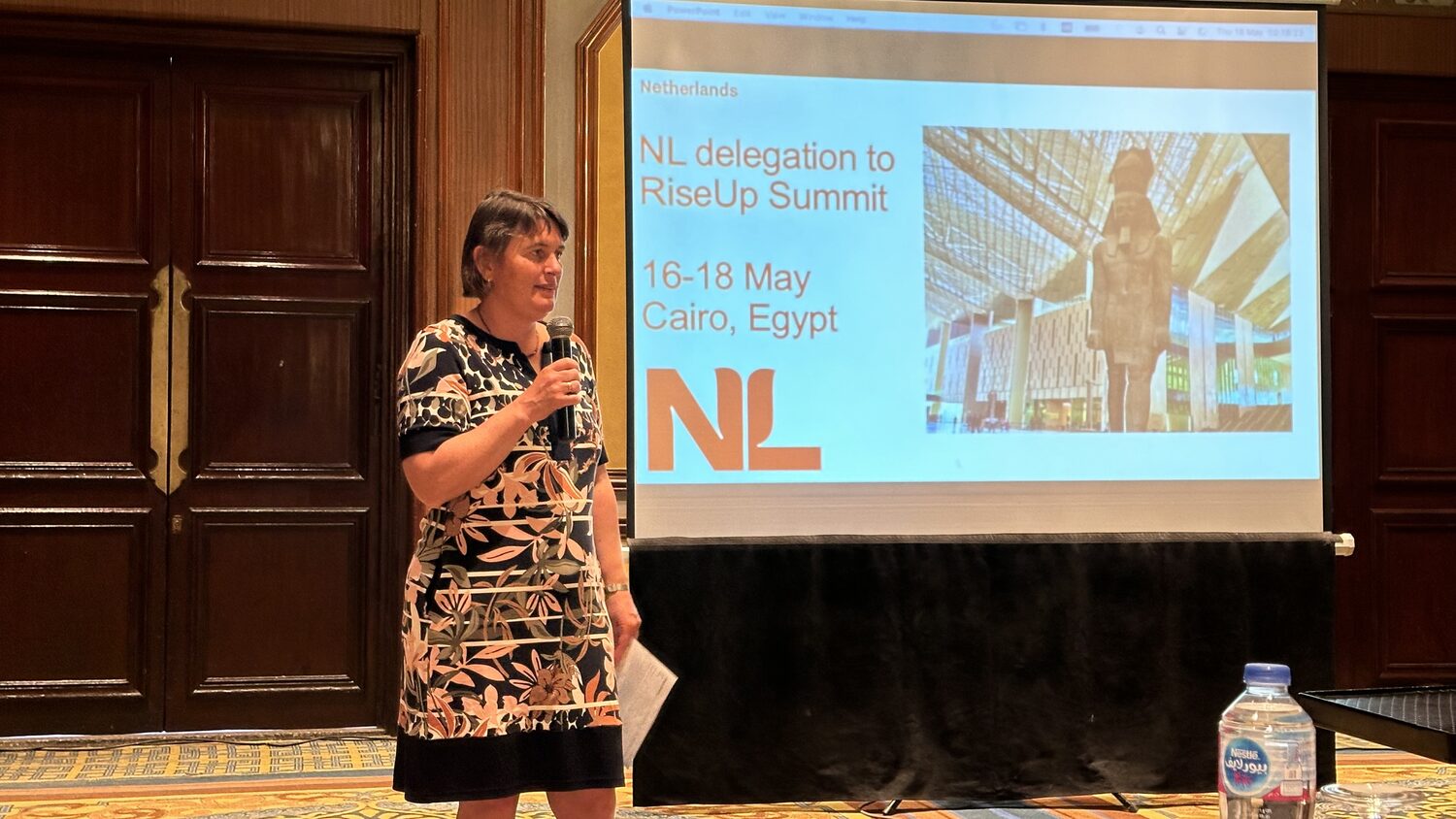 Speaker with a microphone beside a projected slide reading NL delegation to RiseUp Summit, 16-18 May, Cairo, Egypt