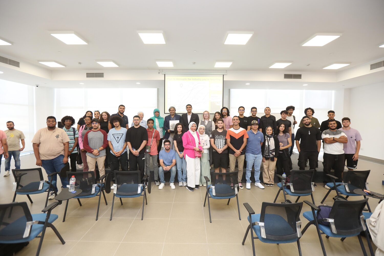 The full student cohort standing together in the EUE lecture room, chairs pushed back, a line chart on the projection screen behind them; Tarry Singh at the left holding a presentation plate in its case.