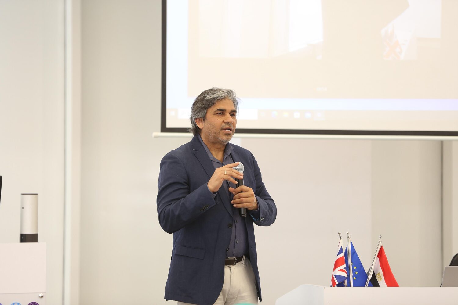 Tarry Singh speaking with a hand mic in front of the projection screen, British, European and Egyptian flags standing on the front desk to his right.