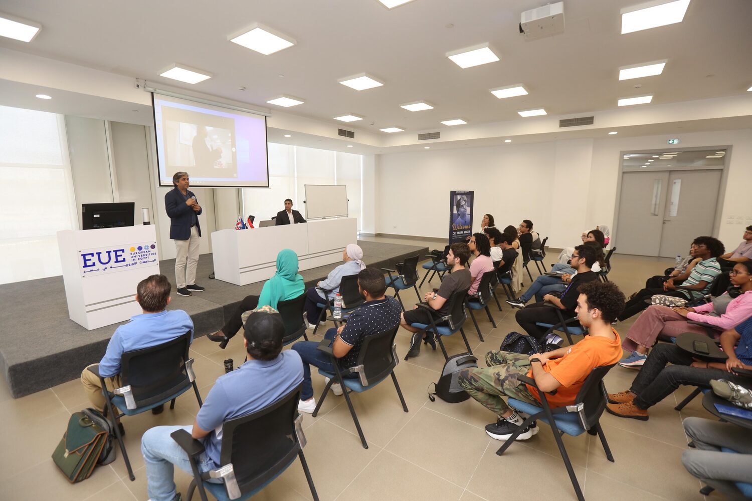 The lecture room seen from the back, students seated across the floor, the EUE lectern at the front and a welcome poster against the side wall.