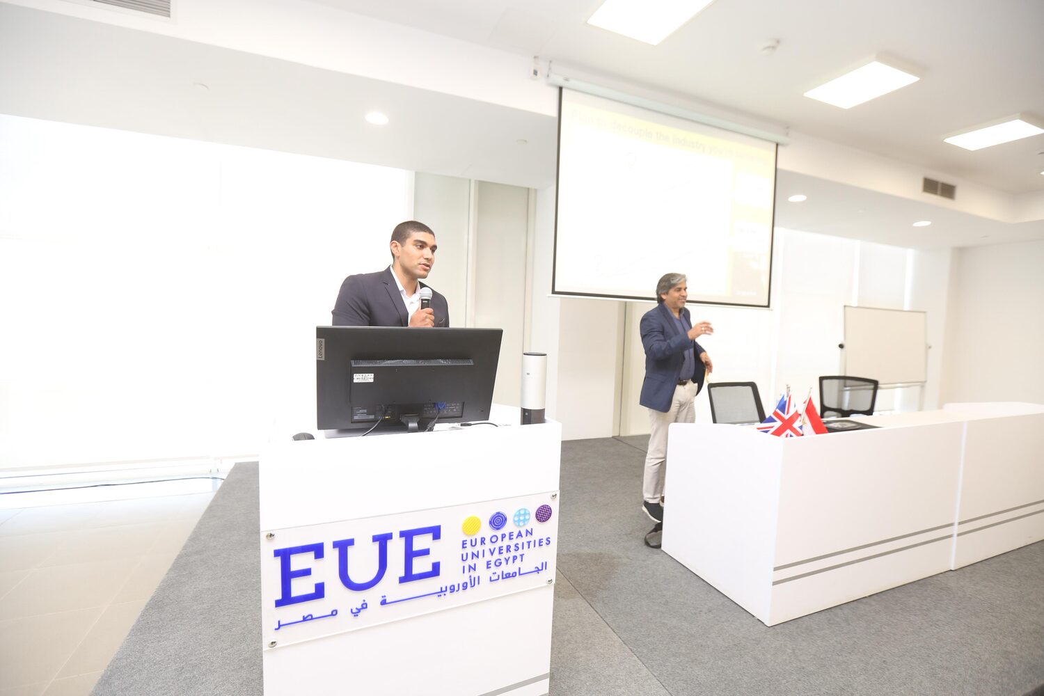 A host addressing the room from the EUE lectern while Tarry Singh stands at the front desk beside the flags.
