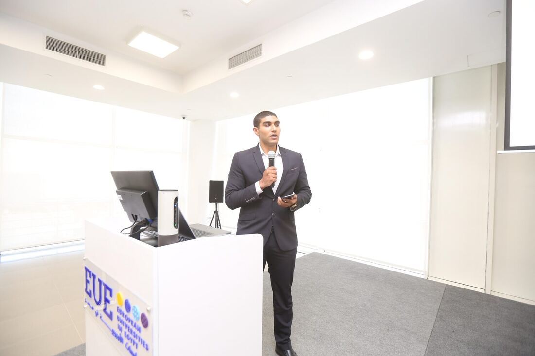 A host speaking into a hand mic beside the EUE lectern in the lecture room.