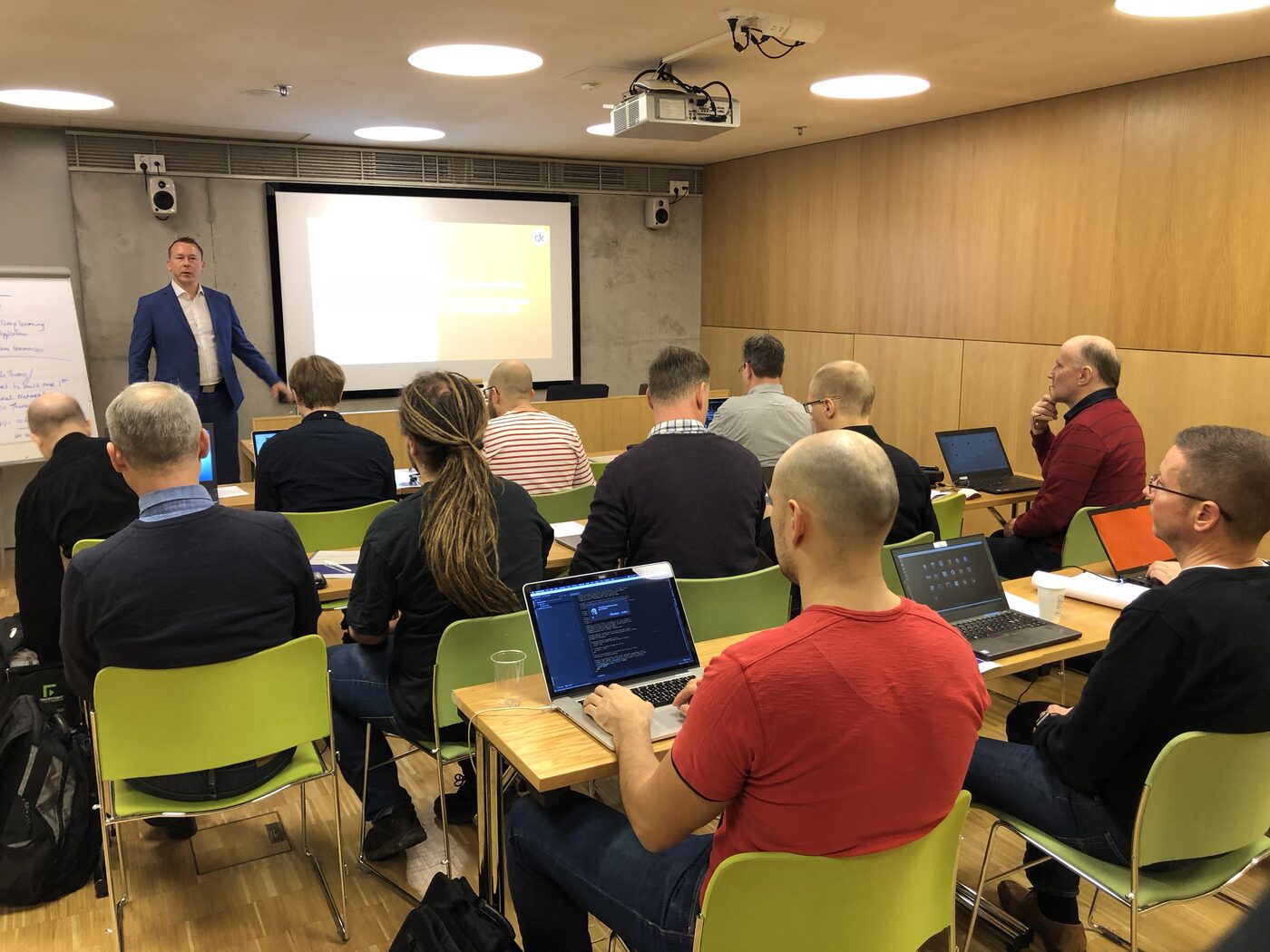 The workshop room in Helsinki during a session, participants seated at desks with laptops open on code, a projected slide on the screen and a flipchart at the front.