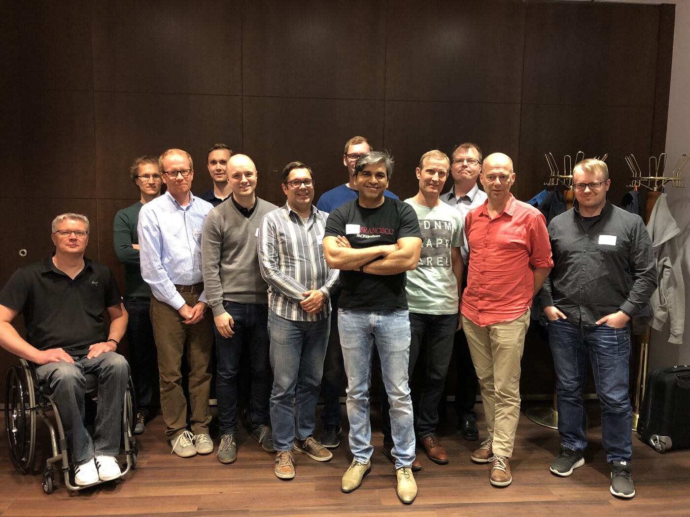 Closing group photograph of the Helsinki cohort, twelve people including Tarry Singh against a wood-panelled wall, several wearing Ari Hovi name badges.