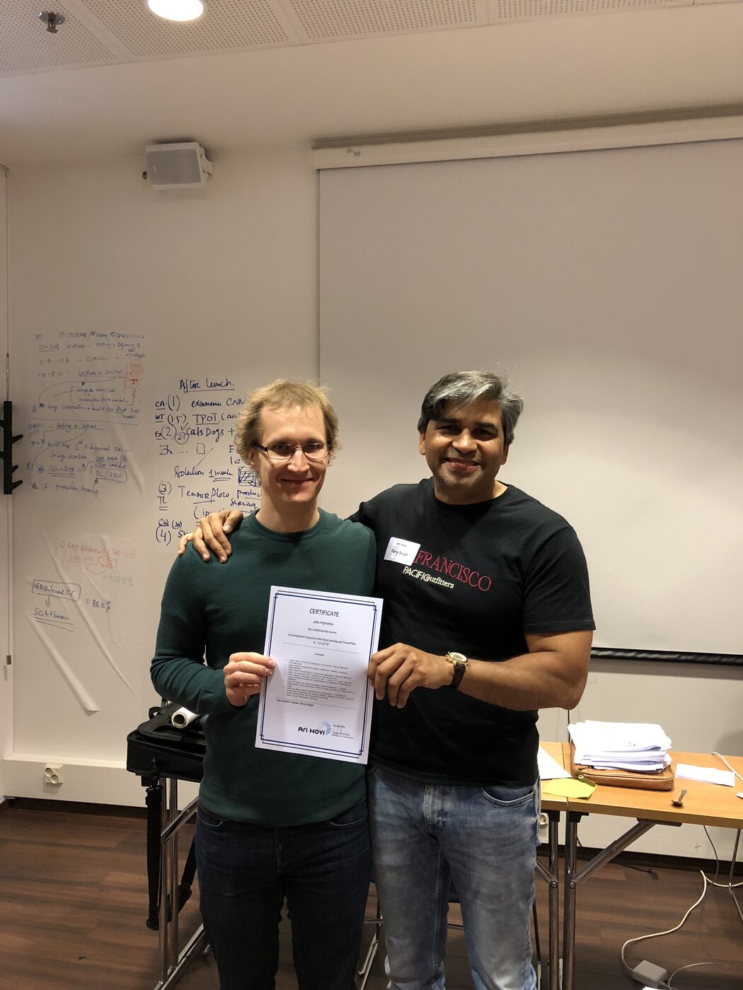 Tarry Singh hands a course certificate to a participant. The certificate reads AI development in practice with Deep Learning and TensorFlow, 6.-7.6.2018, above the Ari Hovi Oy mark, with the two-day agenda whiteboard behind them.
