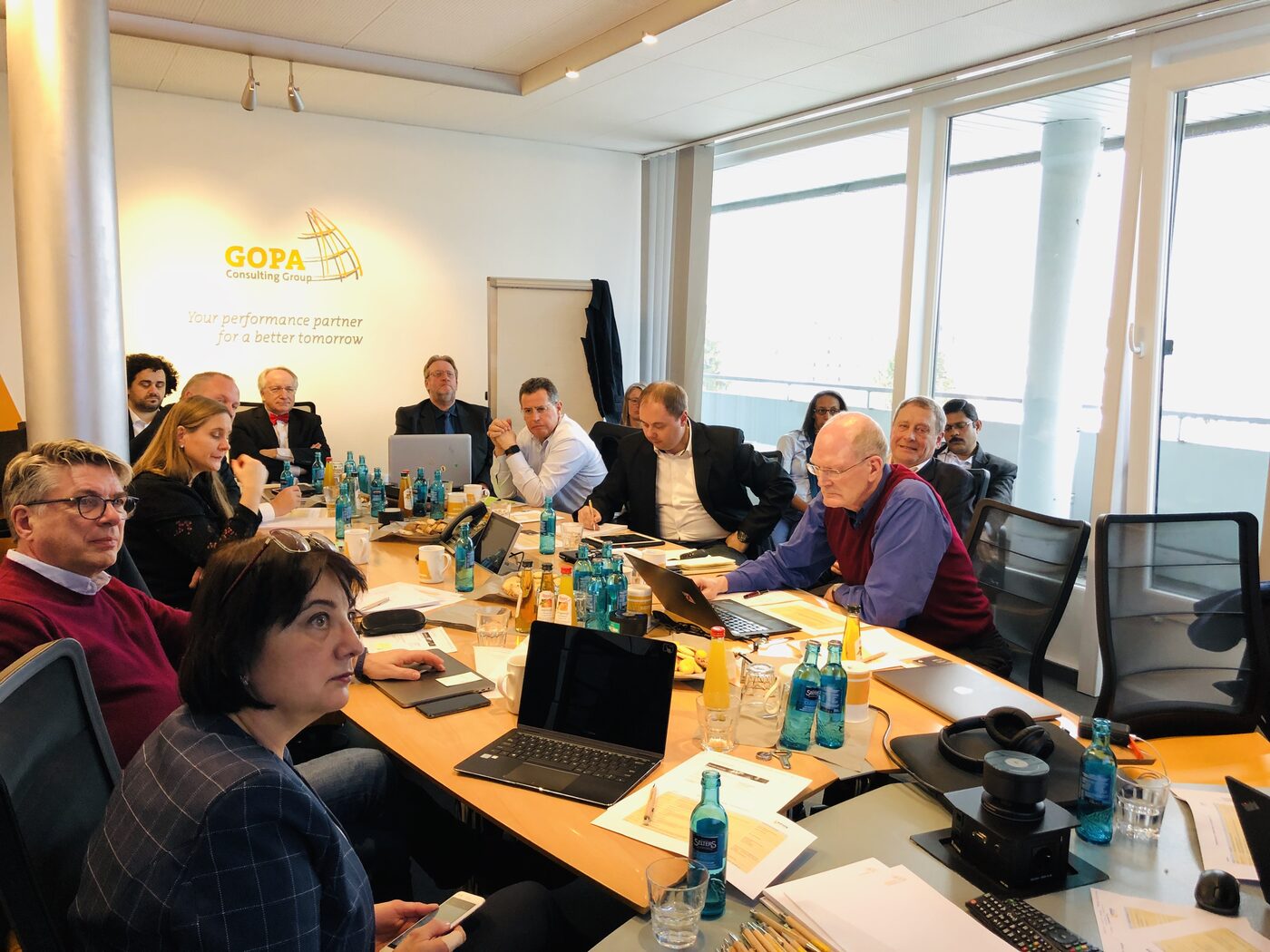 Participants seated around a long conference table at the GOPA Consulting Group offices, with a GOPA roll-up banner against the window wall behind them and laptops and papers across the table.