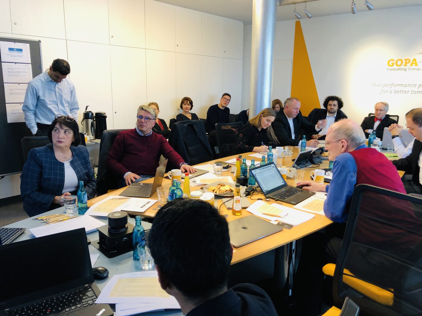 The same meeting room seen from the opposite end, the table full of participants, a GOPA banner on the wall behind and a standing colleague at the left.