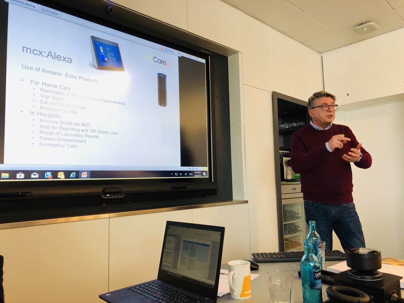 A speaker in a maroon jumper presenting beside the wall screen, which shows a slide on the use of voice assistant devices in home care and in hospitals.