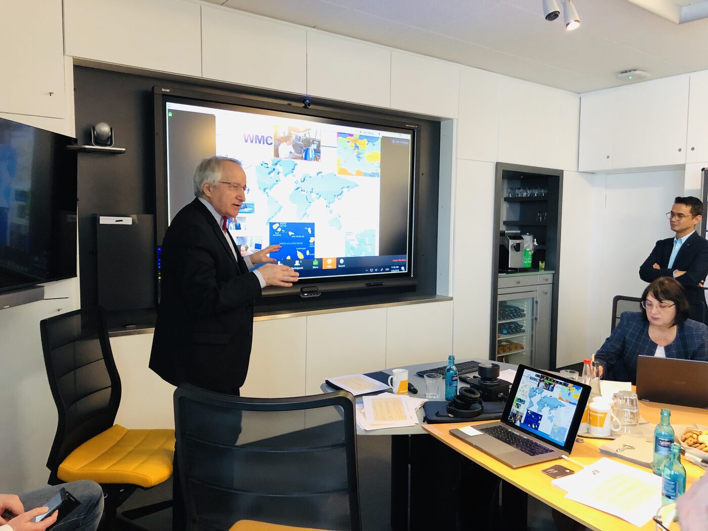 A speaker standing at the wall screen presenting a world map slide to the seated room, with participants at the table and against the back wall.