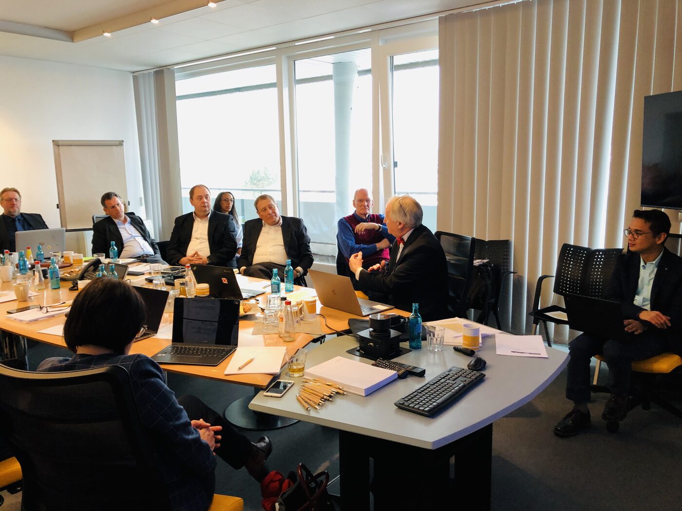 Participants around the conference table mid-discussion, laptops and bottled water on the table, window wall and a flip chart behind.