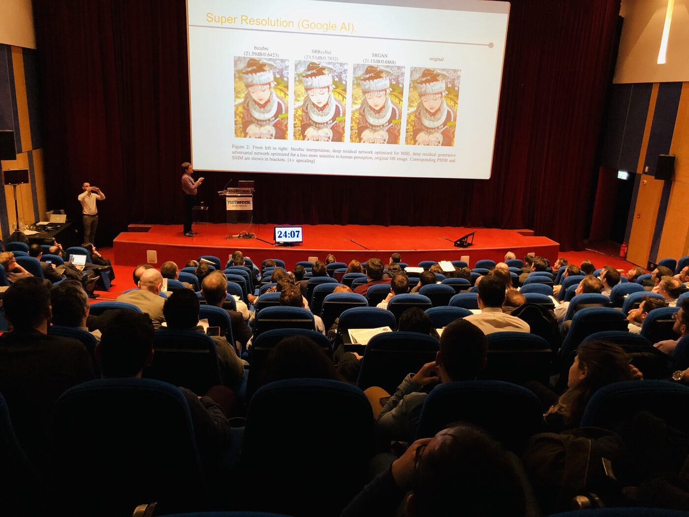 The auditorium mid-session at the Data Conference 2019. A speaker stands at the netweek conferences lectern on the red stage, a countdown clock reads 24:07, the screen shows a super resolution comparison, and every visible seat is taken.