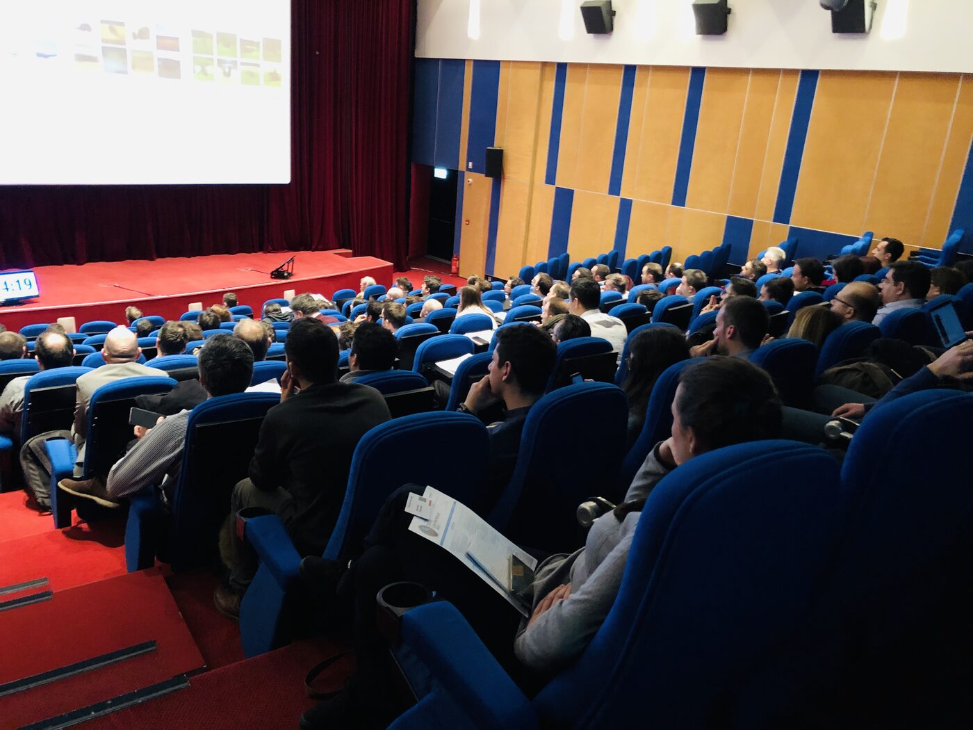 The audience seen from the back rows of the raked auditorium, blue seats filled, the lit screen and red stage ahead and a countdown clock at the left of the stage.