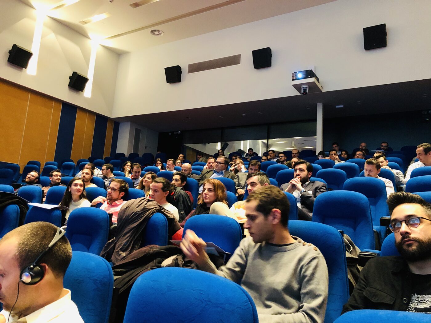 Attendees in the blue seats of the auditorium, several wearing simultaneous translation headsets, watching the stage.