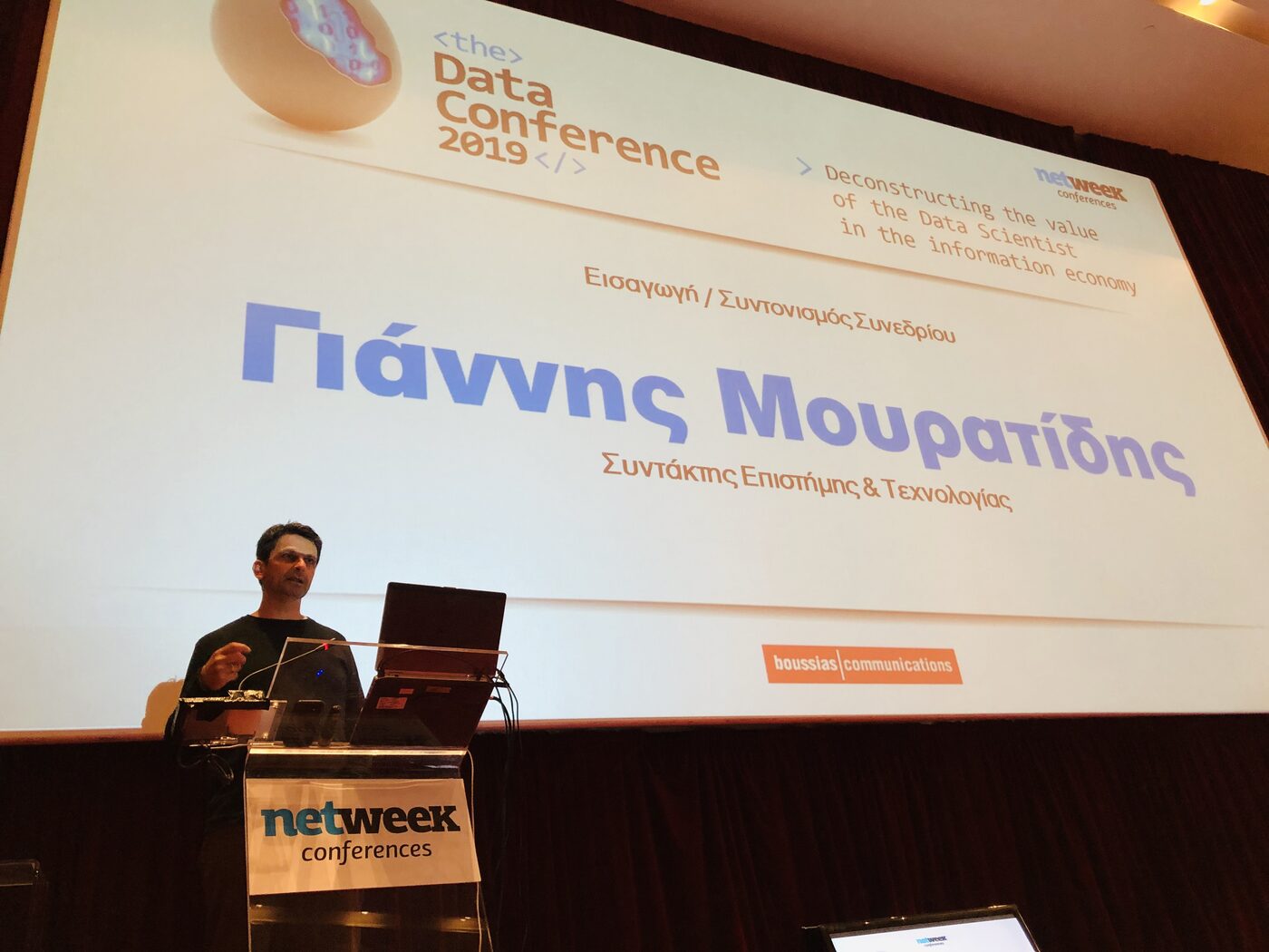 Yiannis Mouratidis opening the conference at the netweek conferences lectern, with the title slide behind him reading the Data Conference 2019 and its strapline on deconstructing the value of the data scientist.