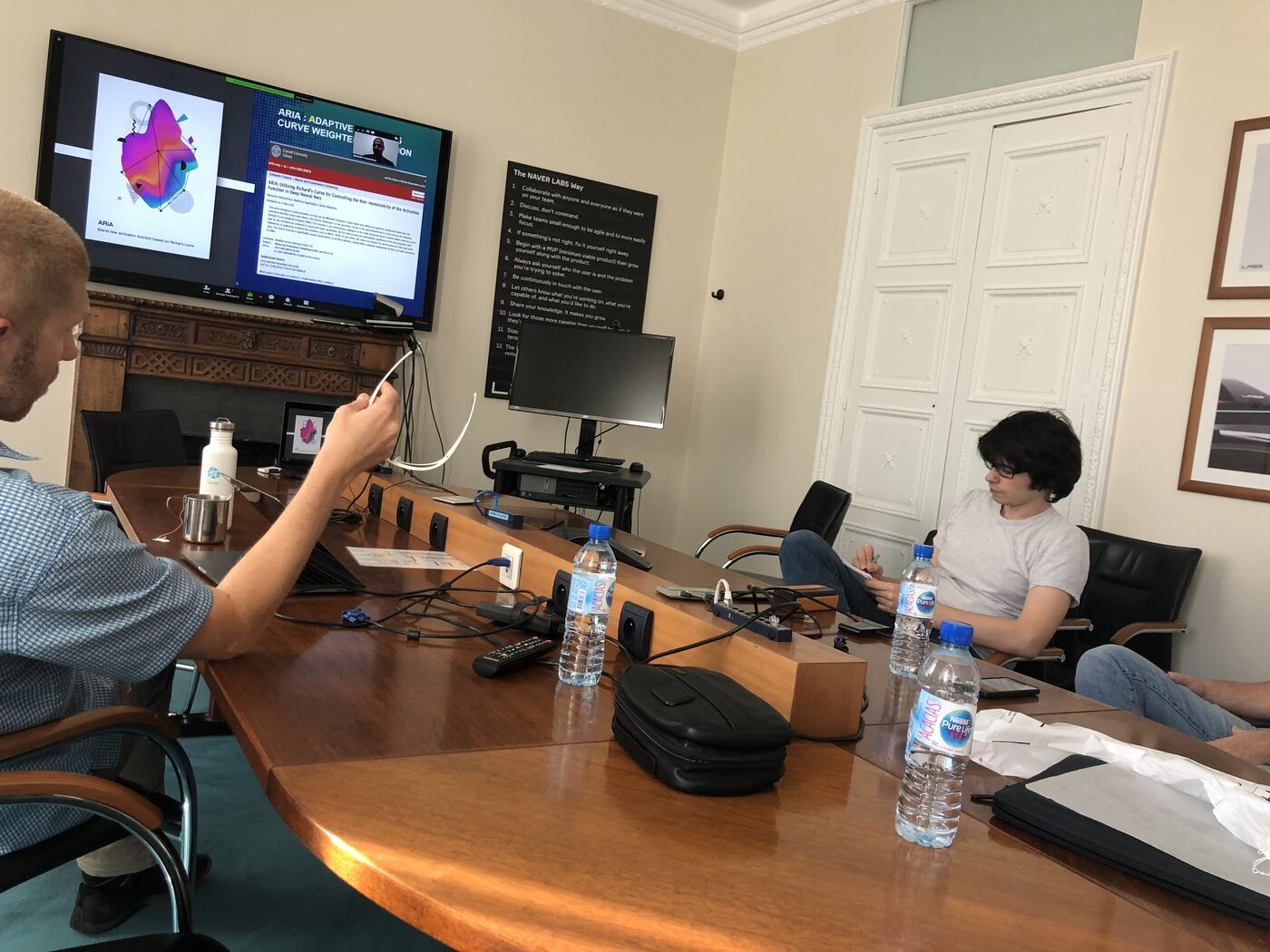A NAVER Labs Europe meeting room during the session. The wall screen carries the ARiA title slide alongside the arXiv listing for the paper, with co-author Narendra Patwardhan joining by video call. A printed copy of The NAVER LABS Way hangs on the wall to the right.