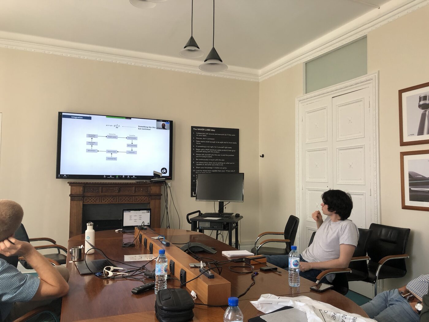 The LAYGo integration slide on the wall screen, showing the fuzzy-logic controller flow diagram, The NAVER LABS Way poster alongside and a participant seated at the table.