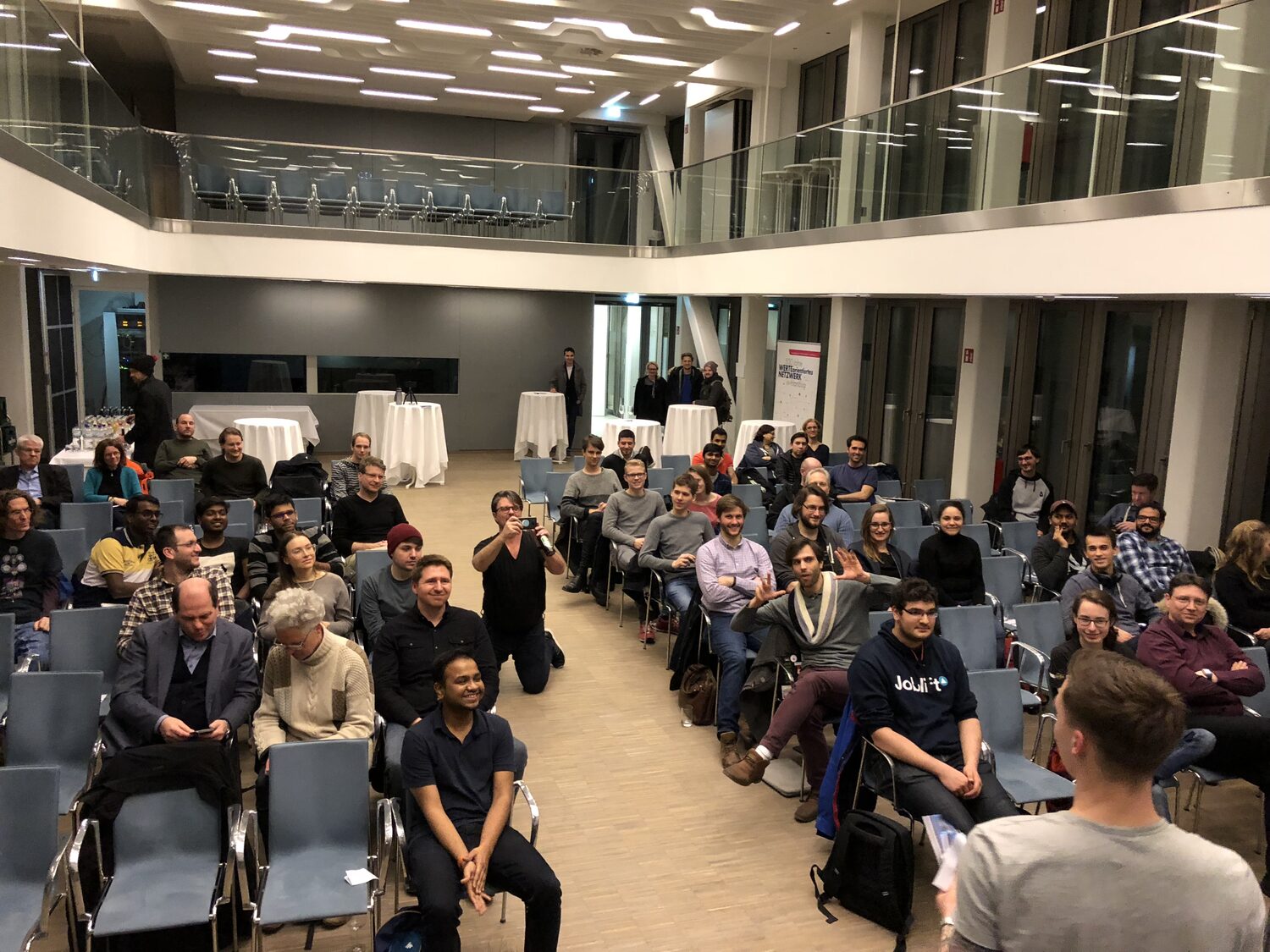 The HSBA atrium hall seen from the back at 20:57, around fifty people in blue chairs under a first-floor gallery, drinks tables and a pull-up banner behind them