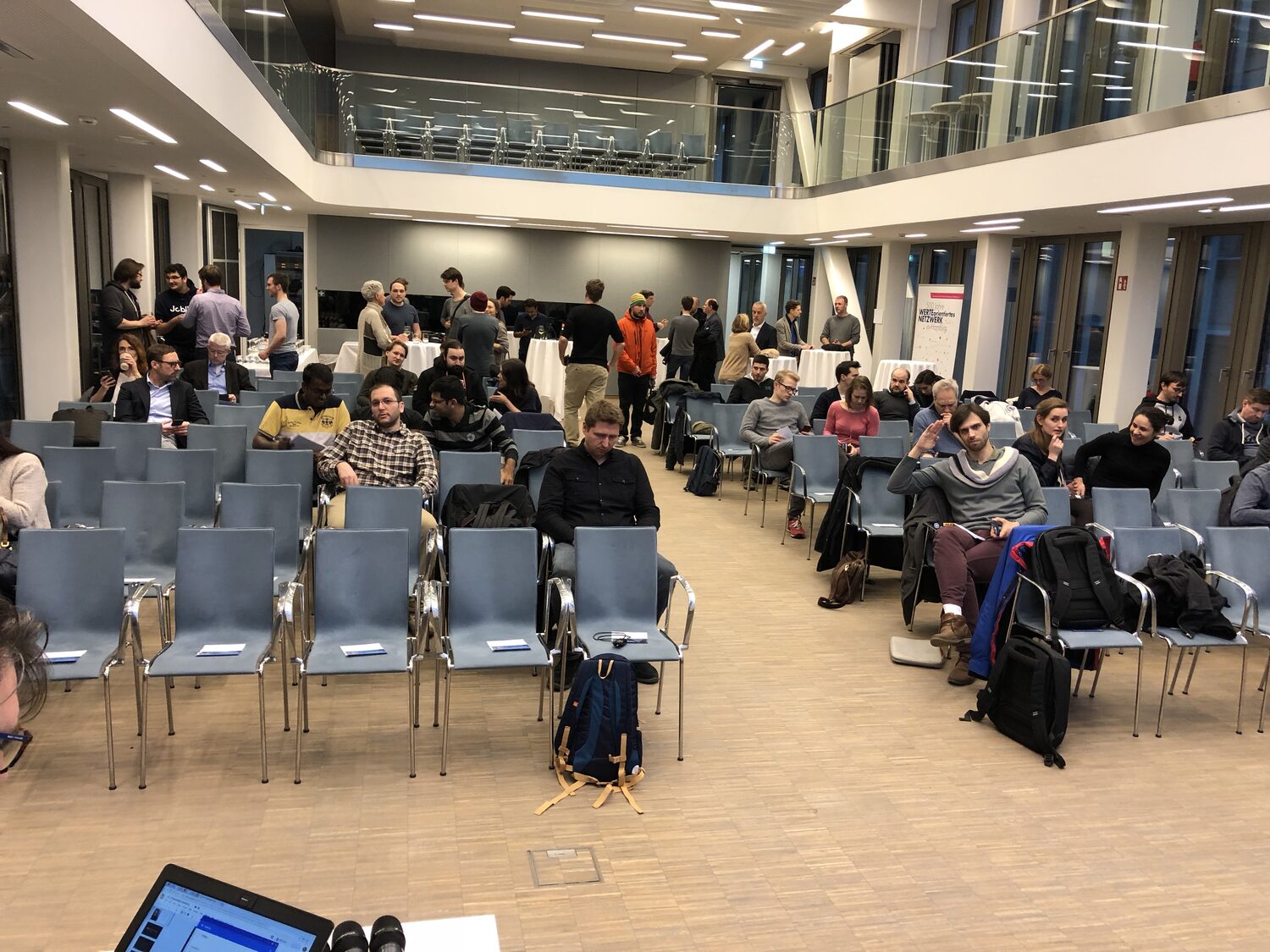 The hall filling up at 18:20, rows of blue chairs seen from the side, camera and laptop on the tech desk in the foreground