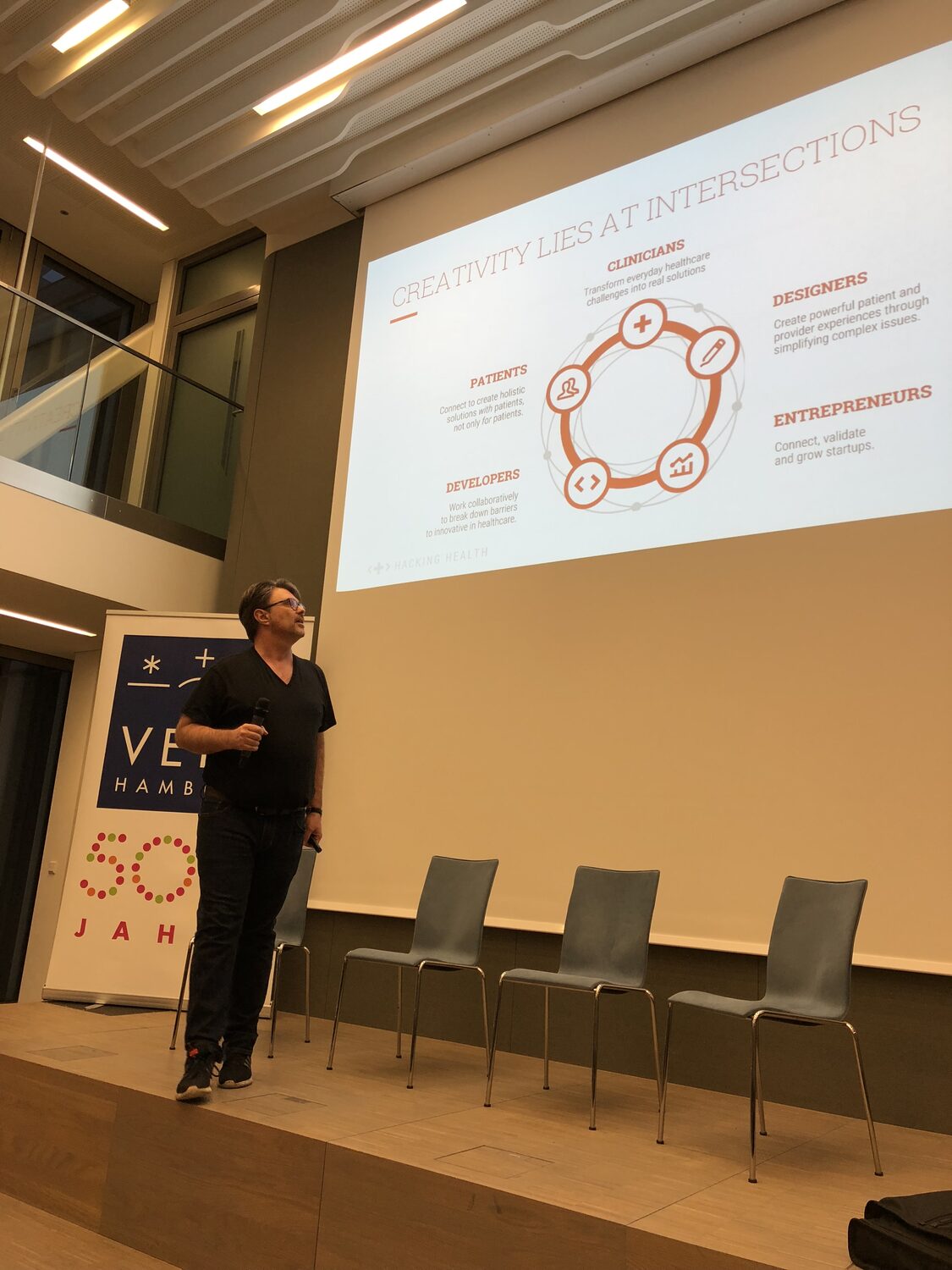 A speaker on stage with a Hacking Health slide reading Creativity lies at intersections, beside a navy pull-up banner marked Hamburg and 500 Jahre