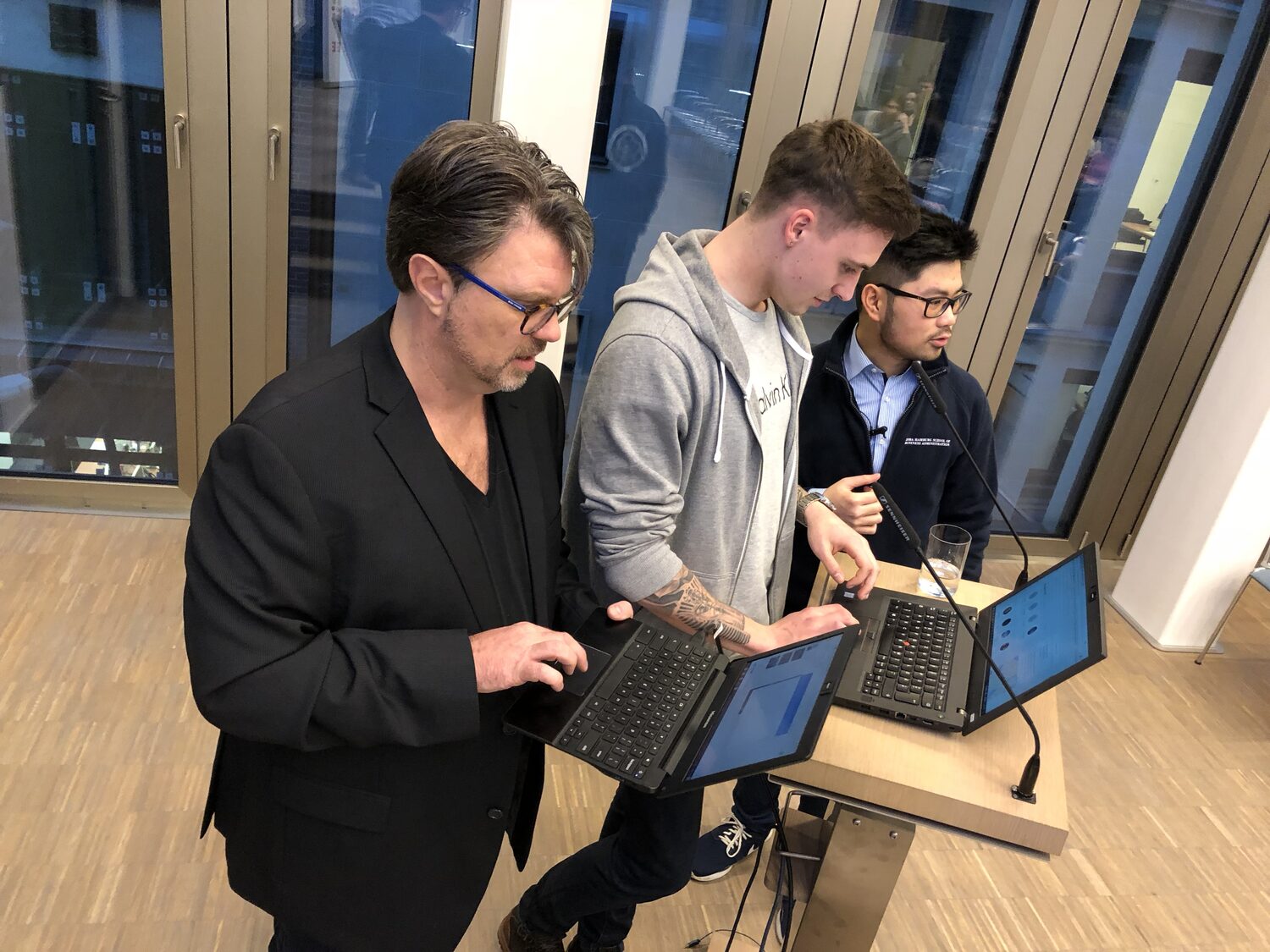 Three people at the lectern trading laptops before the programme, one wearing an HSBA Hamburg School of Business Administration fleece and holding a handheld microphone