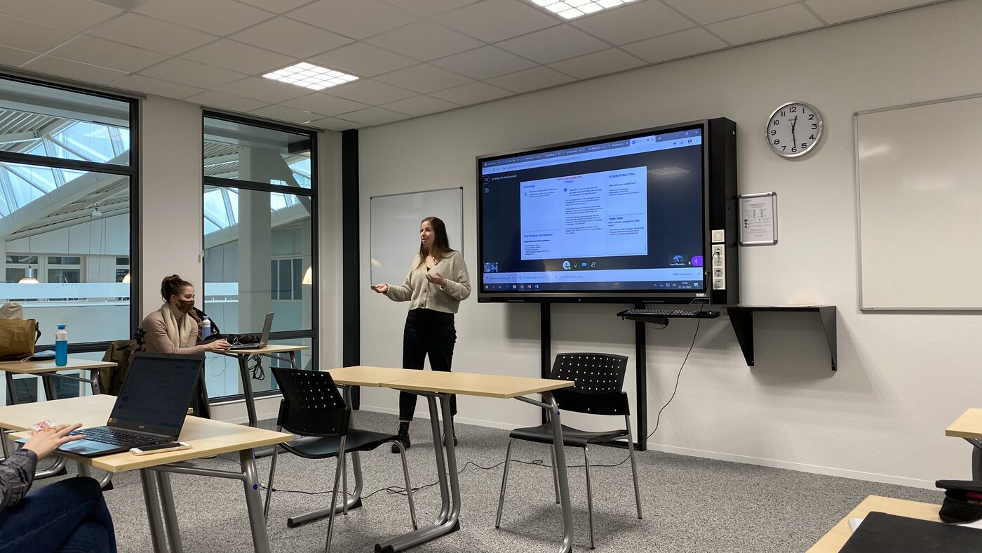 A student presents beside a wall-mounted display running a shared browser session. A masked classmate works at a laptop by the window. The wall clock reads just past four.