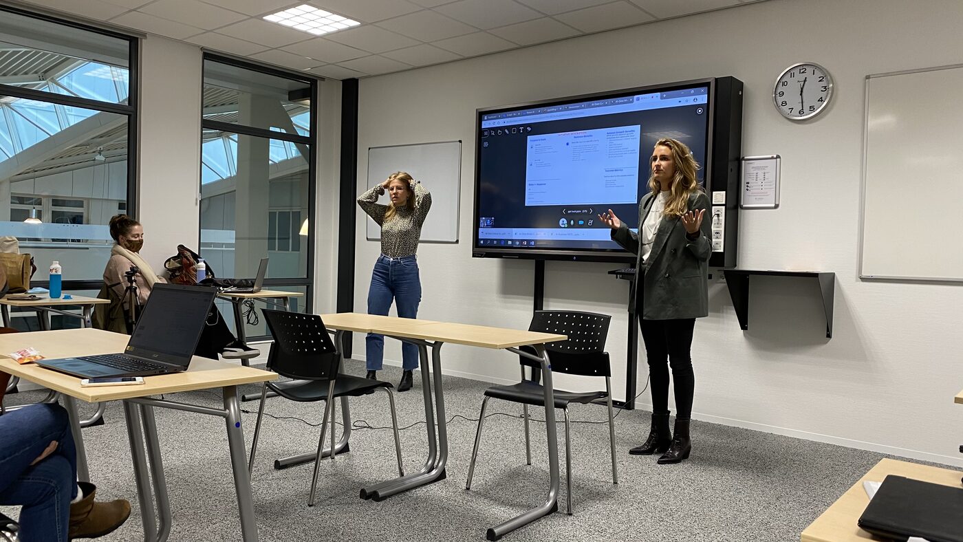 Two students stand either side of the display while a third shares an initiative description canvas from a remote connection.