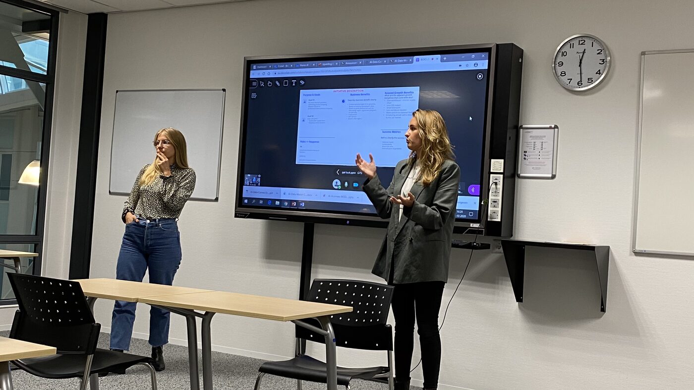 A pair of students take questions in front of the display, the initiative description canvas still on screen.
