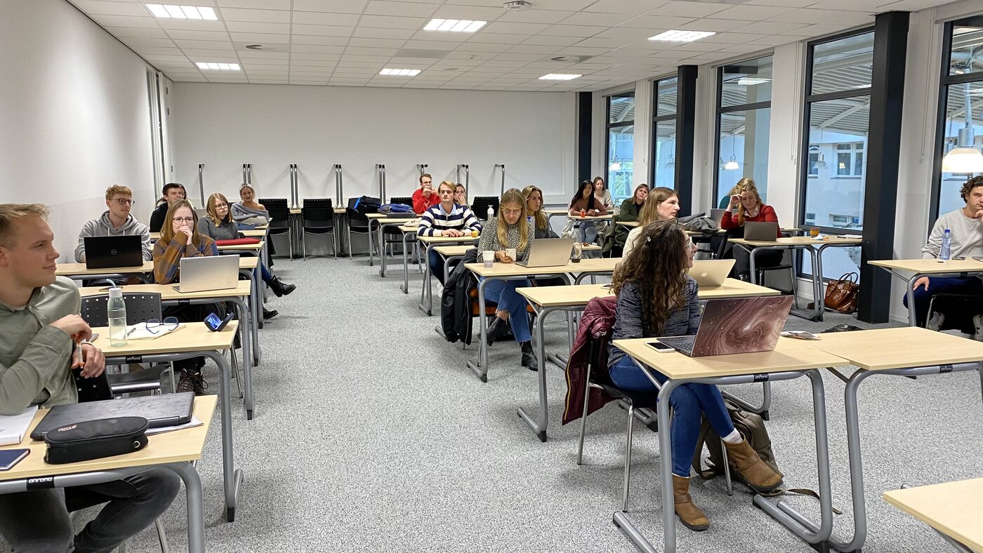 The room from the front corner. Rows of separated desks, an empty block at the back, students turned toward the display.