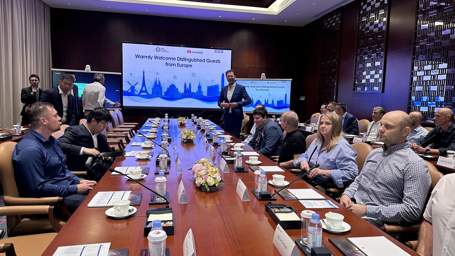 Huawei boardroom at the Bantian base, Shenzhen, 30 June. A delegate stands and speaks beneath a screen reading Warmly Welcome Distinguished Guests from Europe, carrying both the Fiber Carrier Association and Huawei marks. Name cards line the long table.