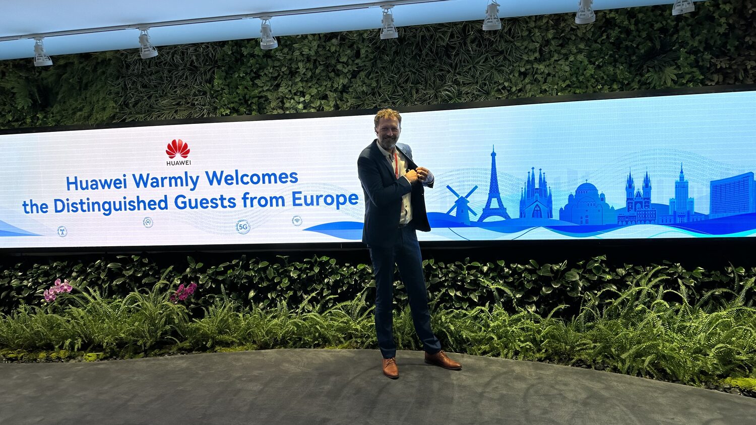 A delegate stands on a low stage in front of a wide LED banner reading Huawei Warmly Welcomes the Distinguished Guests from Europe, backed by a living green wall. Shenzhen, 30 June, late afternoon.