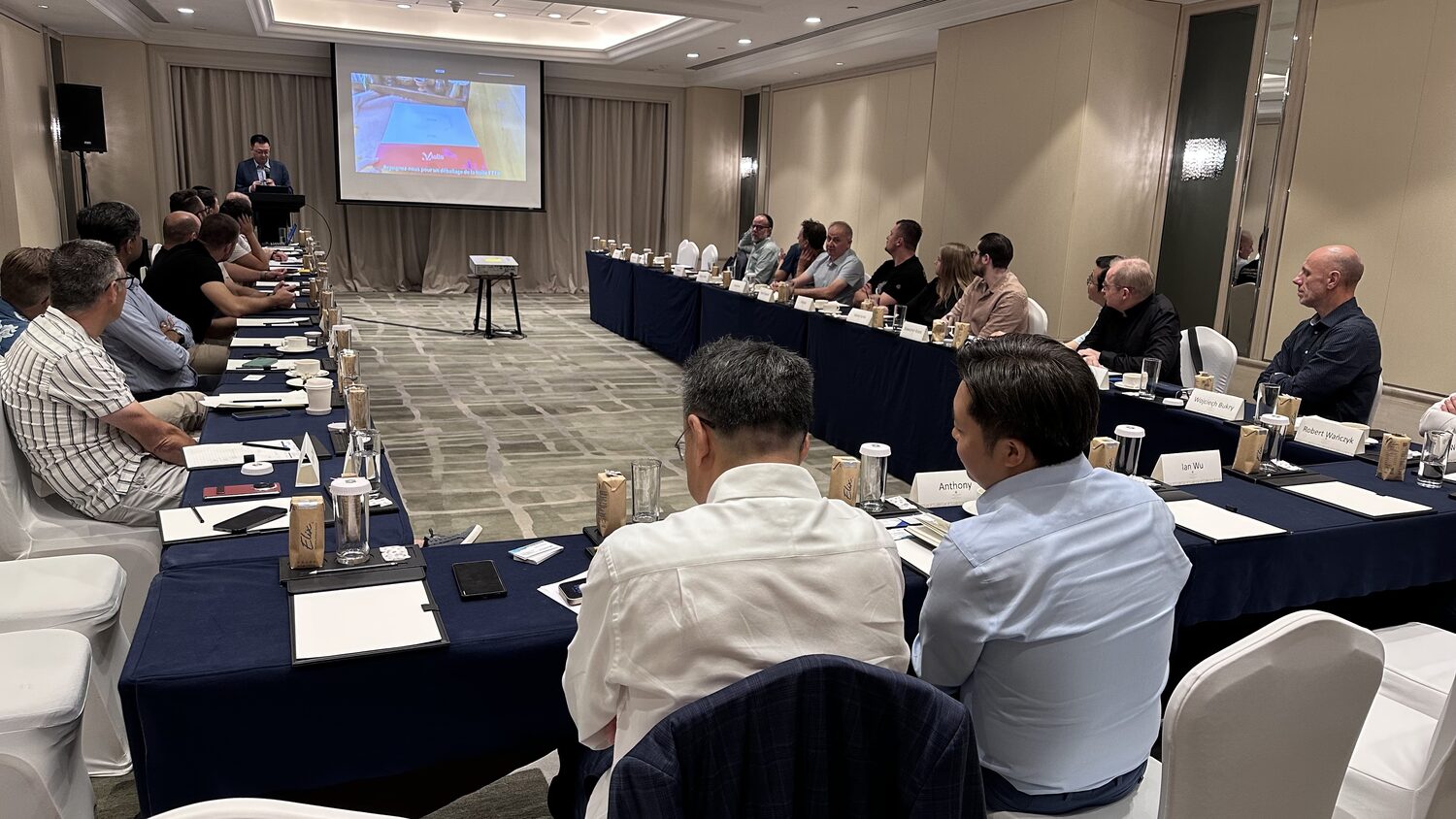 Working session on the final day, Shanghai, 4 July. Tables in a long U with named place cards, a speaker at a lectern, projector on a stand.