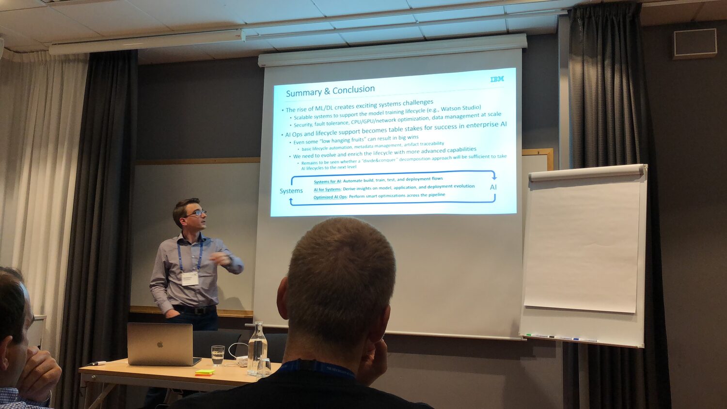 A speaker stands beside a projected slide headed Summary and Conclusion in a small conference room, audience heads in the foreground.