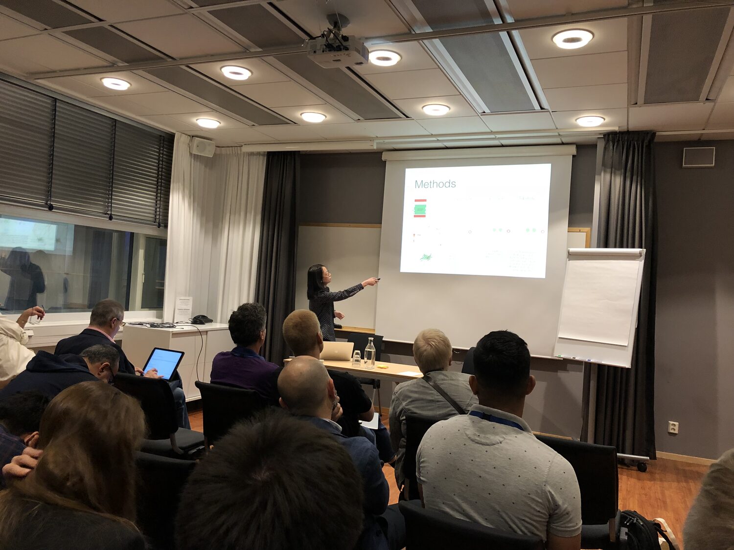 Wide view of a full workshop room, presenter pointing at a slide headed Methods, delegates seated with laptops.