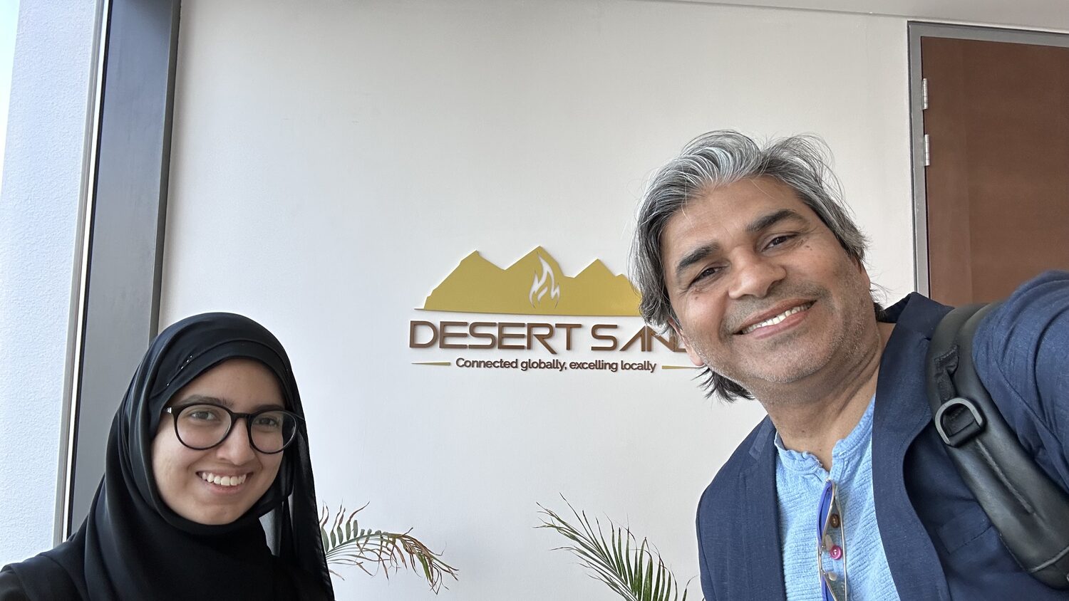 Tarry Singh with a colleague at the Desert Sand Oil & Gas logo wall, under the strapline about being connected globally.