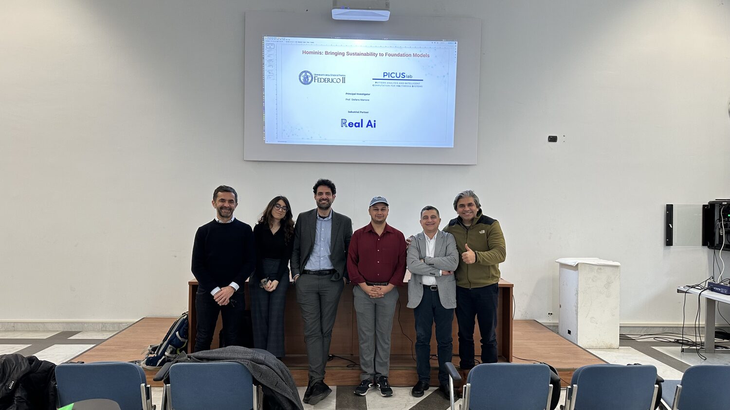 Six people stand in front of the lecture hall dais beneath the projected Hominis title slide carrying the Federico II, PICUS Lab and RealAI logos.