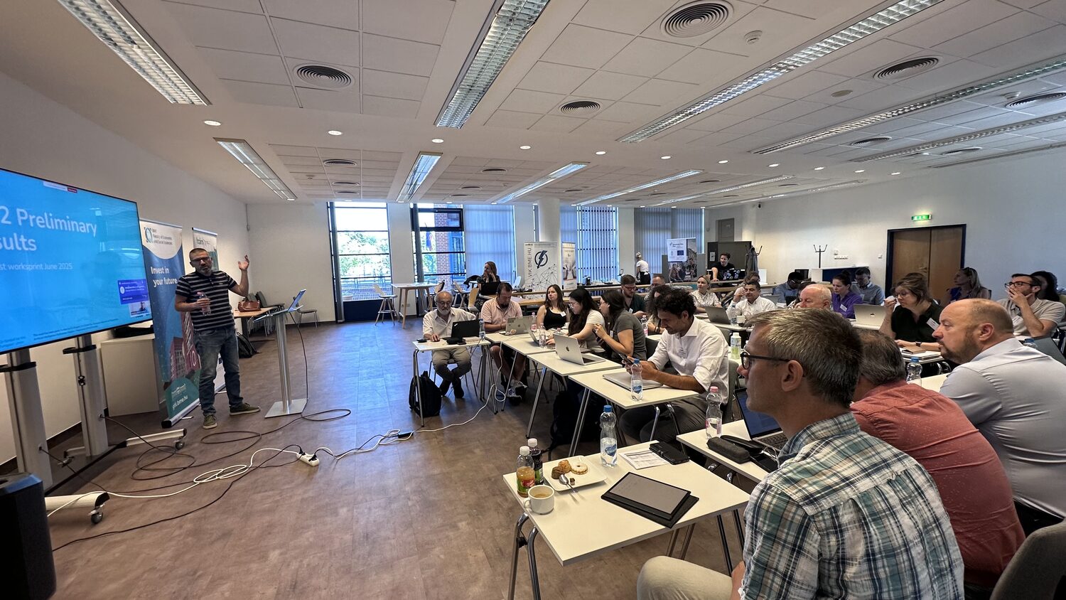 Wide view of the BME meeting room during the PANORAIMA worksprint, a presenter at the screen showing the WP2 preliminary results title slide and partners seated across rows of desks.