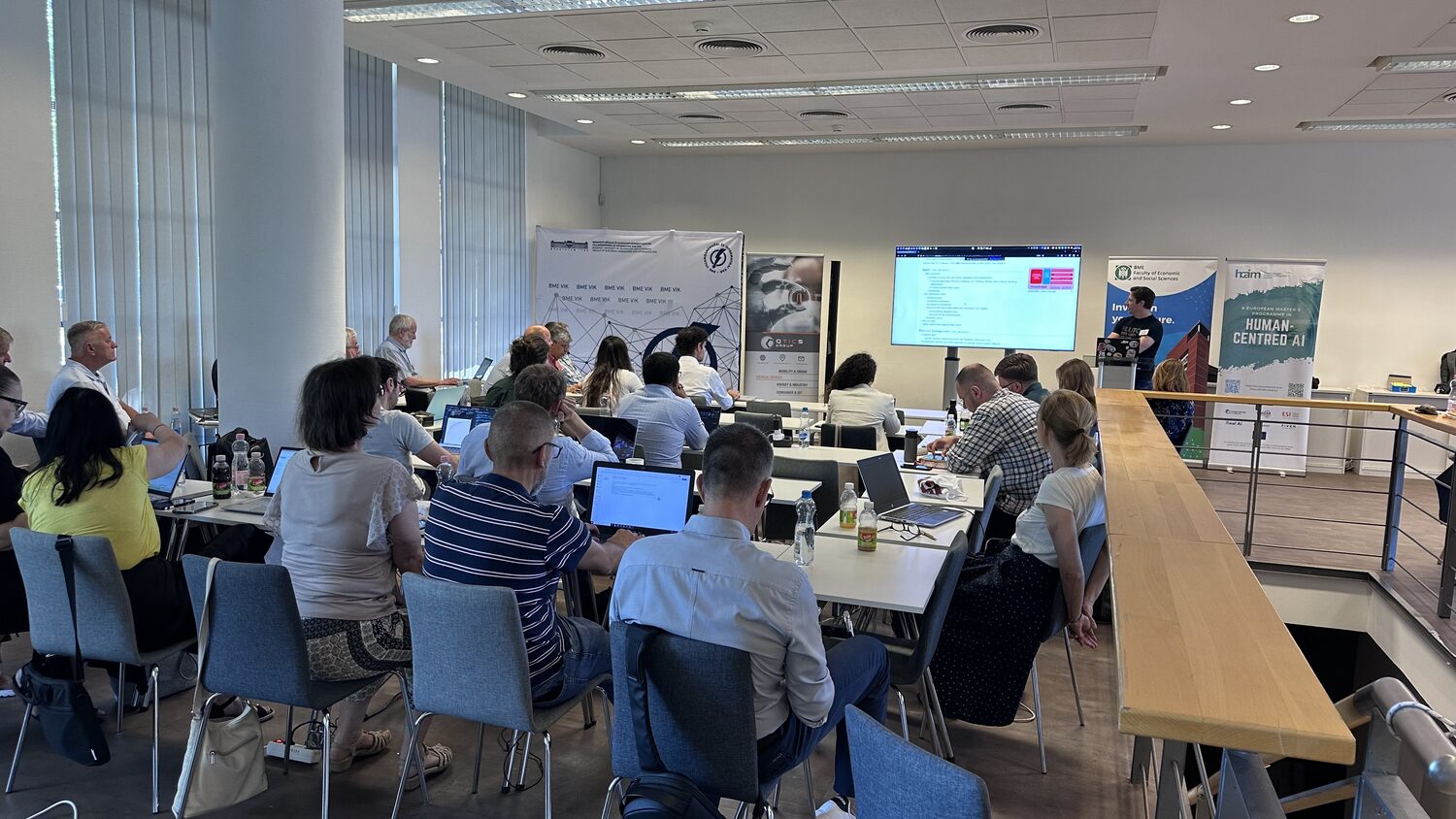 The worksprint room seen from the back, laptops open along the desks and the agenda projected on the far screen between partner banners.