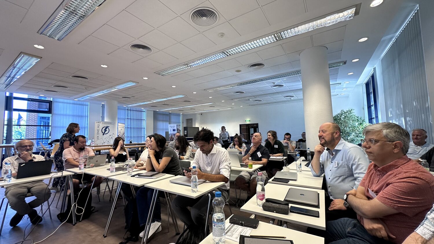 Consortium partners seated at desks in the BME room, listening during a plenary session.