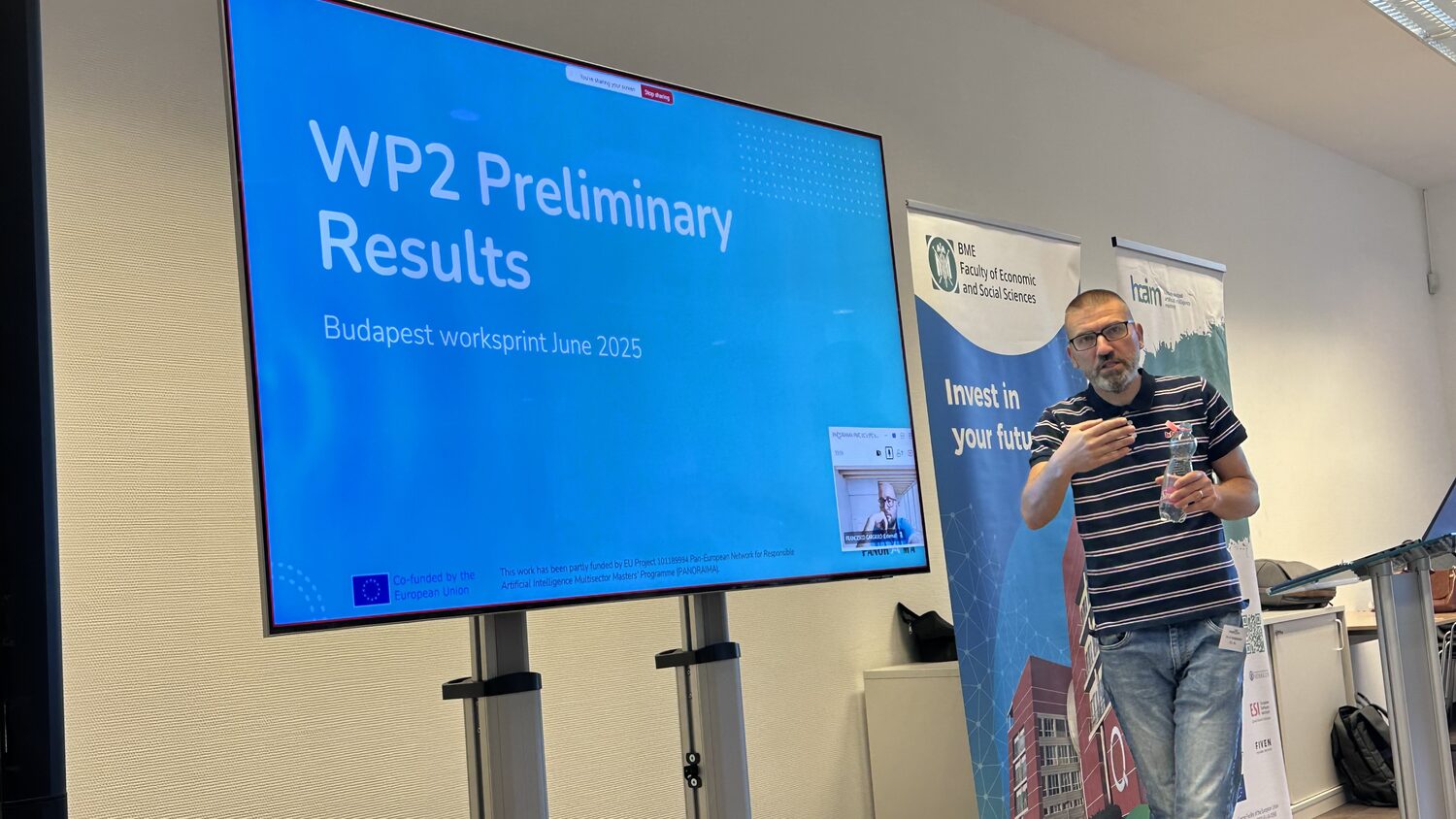 Presenter beside the WP2 Preliminary Results slide, subtitled Budapest worksprint June 2025, with a remote partner in a video window.