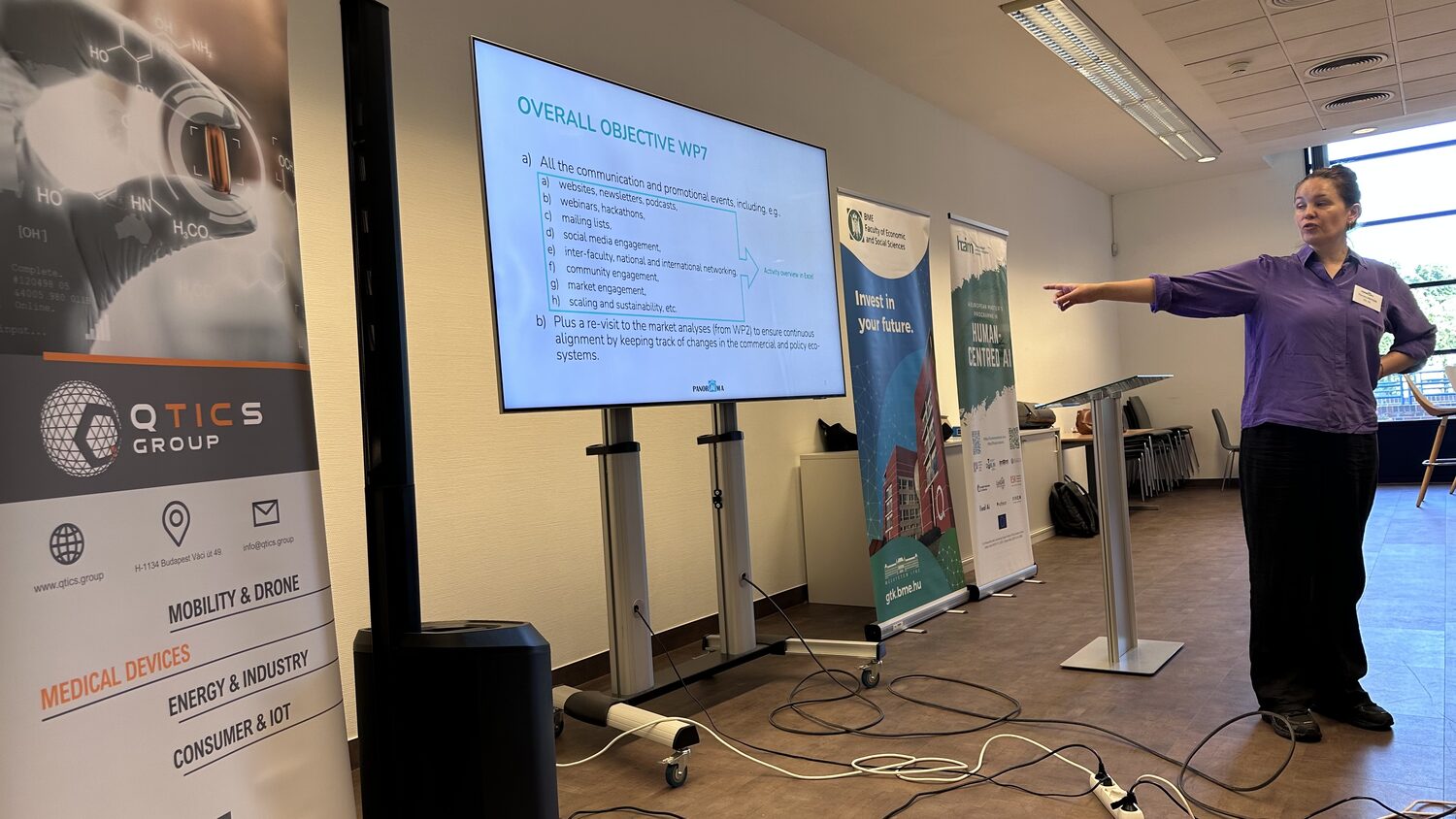 Presenter pointing at the Overall Objective WP7 slide, with the QTICS Group banner in the foreground.