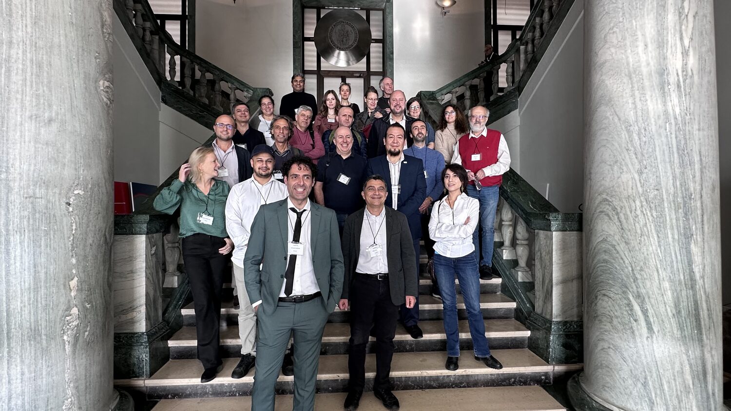 Consortium members grouped on the Federico II staircase between two marble columns, name badges on lanyards.