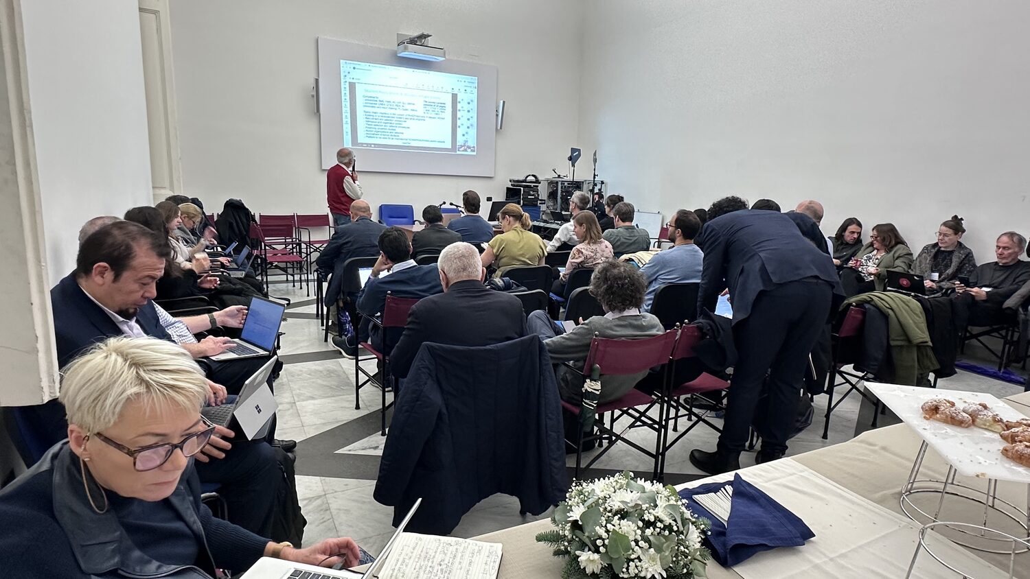 The Naples plenary room seen from the back: delegates at open laptops, the recording rig against the wall, coffee and pastries on a trestle table.