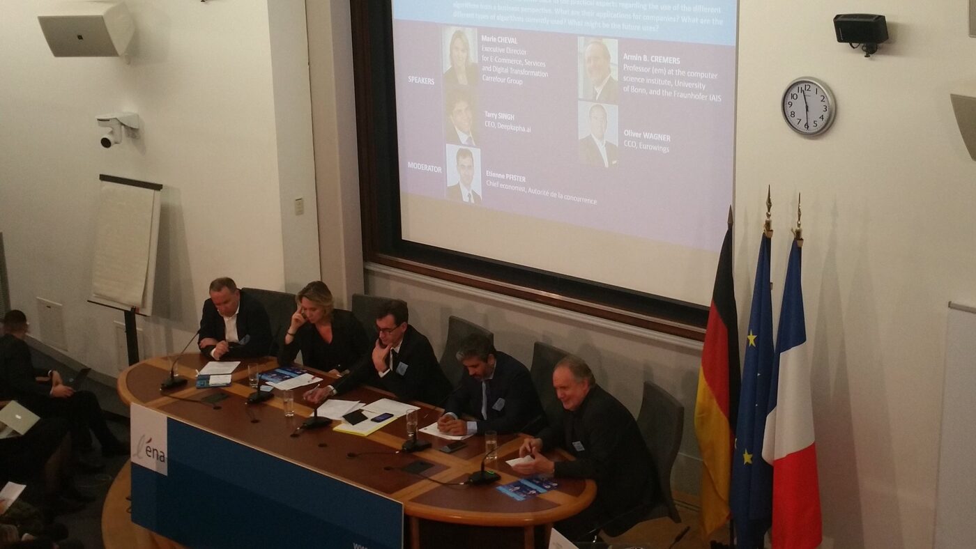 Elevated view of the panel table and part of the audience, with the programme slide listing the four speakers and the moderator legible on the screen.