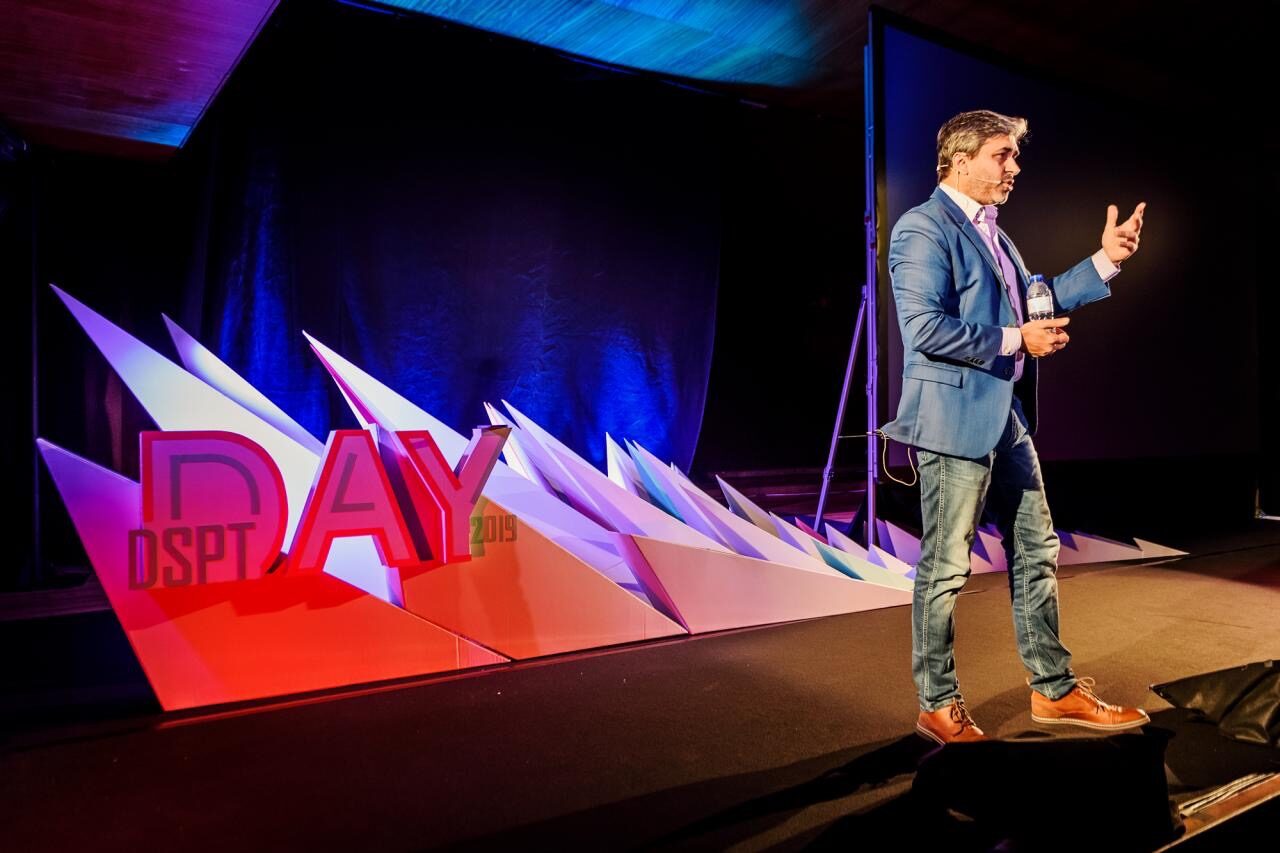 Tarry Singh speaking on the DSPT Day 2019 stage in Porto, gesturing with one hand, beside a lit red and white set piece spelling DSPT DAY 2019.