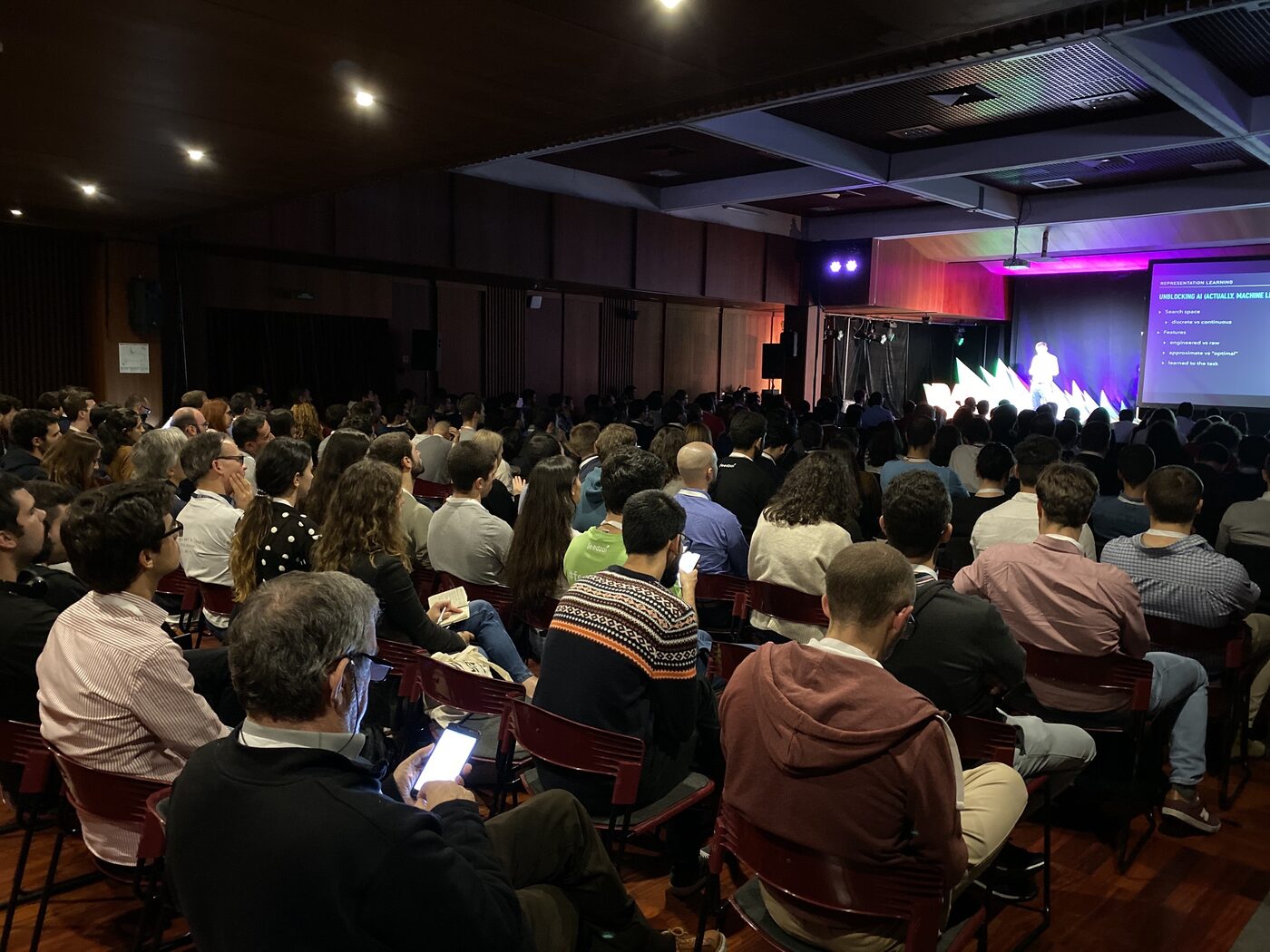 The DSPT Day auditorium seen from the back rows, every seat taken, with a speaker on the distant stage and a slide reading Representation Learning on the screen.