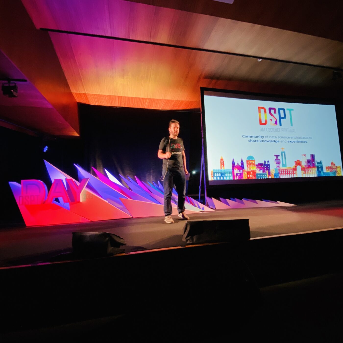 A host in a DSPT Day t-shirt on the Porto stage, the screen behind him showing the Data Science Portugal community slide and a colourful city skyline.