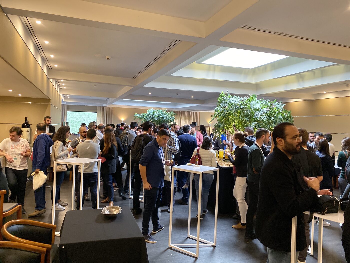 Attendees at the DSPT Day networking break, standing around white high tables in a daylit foyer with a glass roof panel.