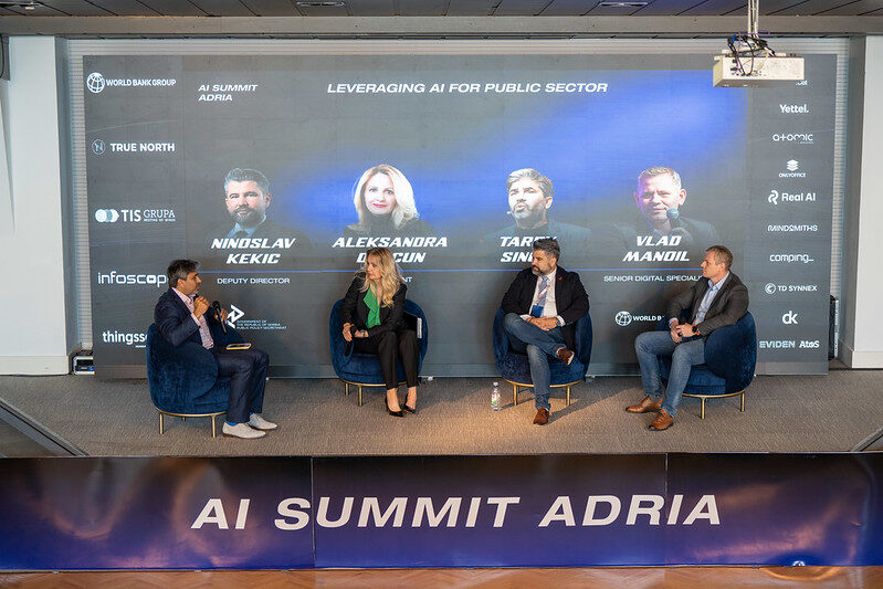 Four panellists in blue armchairs on stage under a screen carrying their names, with an AI Summit Adria banner across the front of the stage.